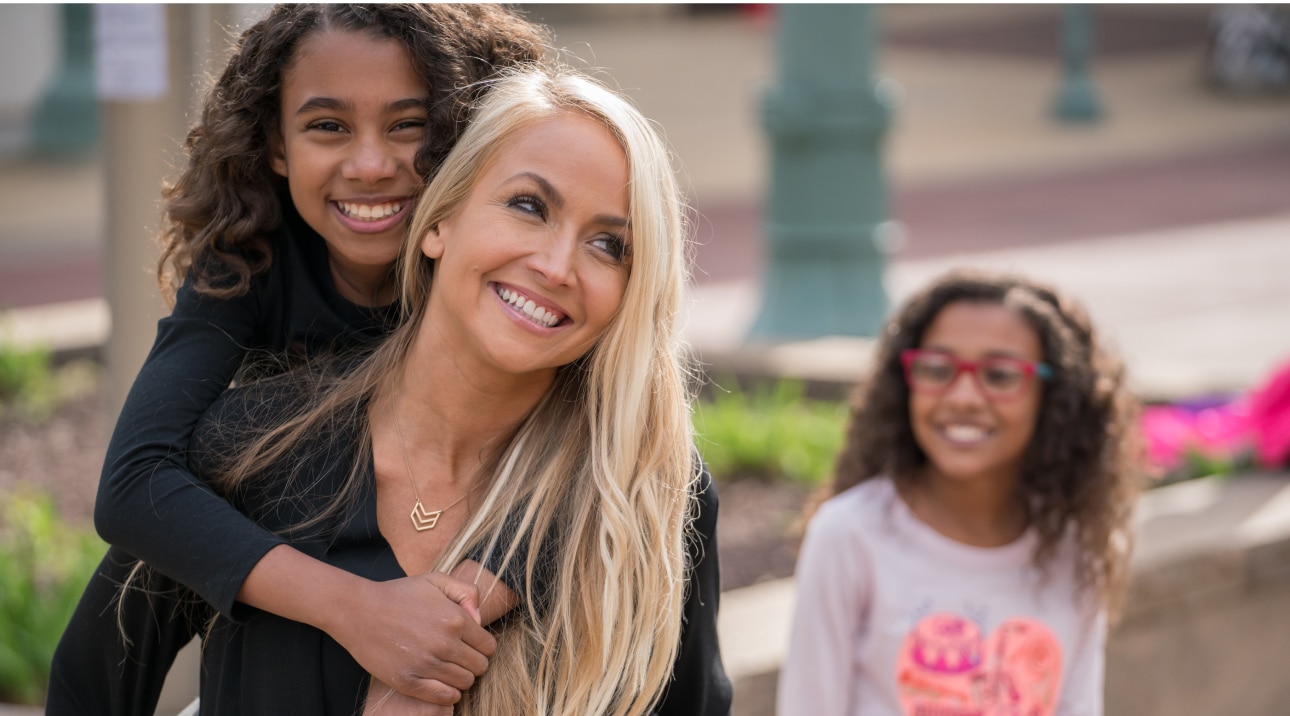 Girl and mother smiling for a picture with younger sister in the background.