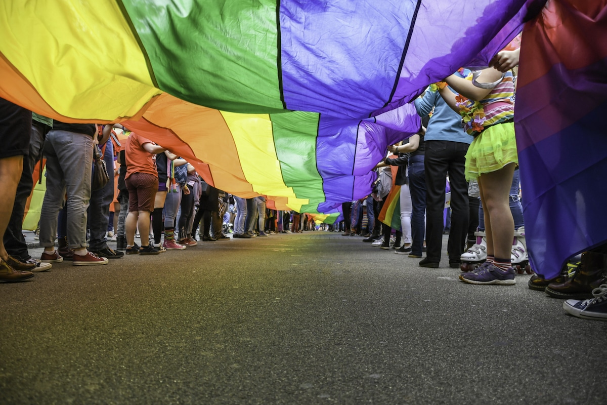 View underneath a long LGBT Pride Flag with rows of people on each side holding it up as it extends down a road