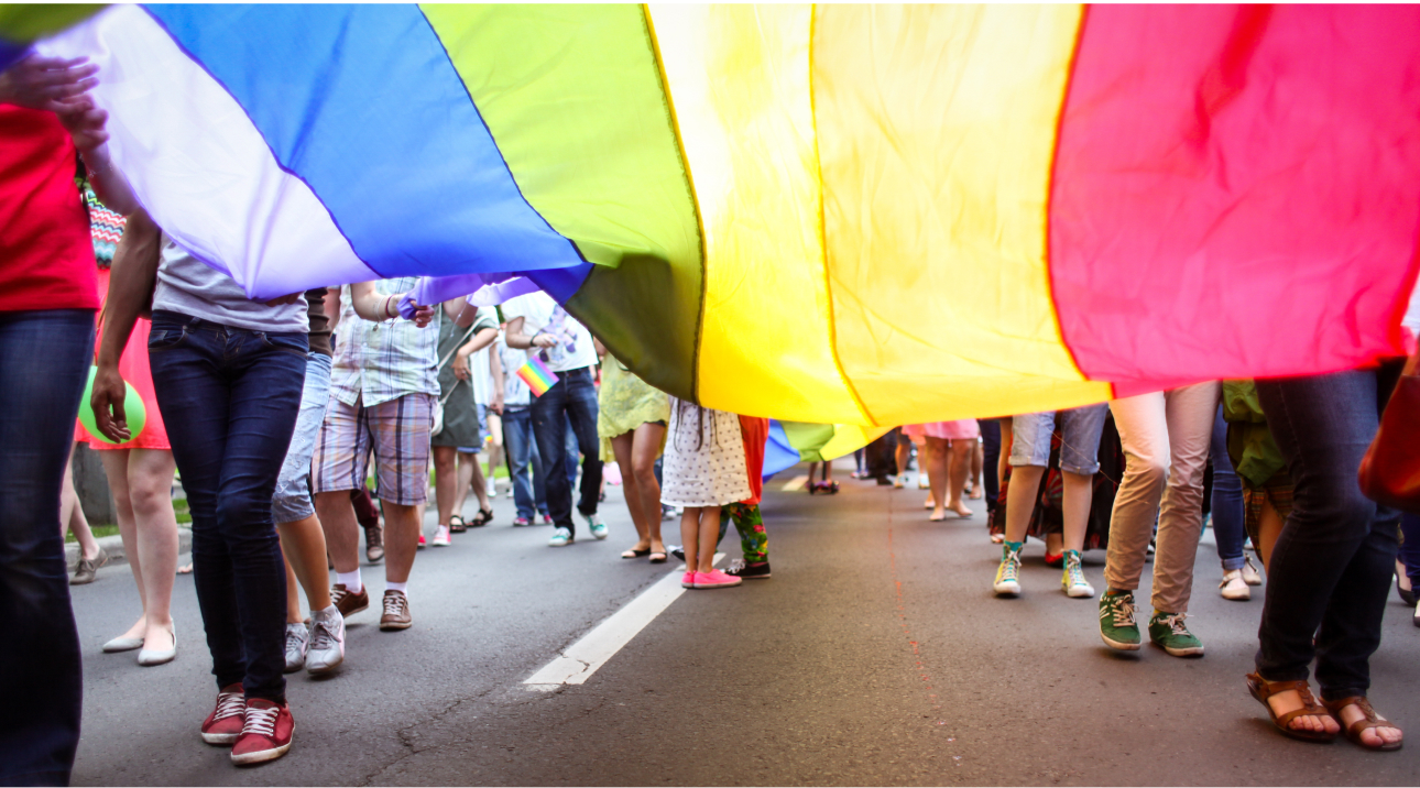 View underneath a long LGBT Pride Flag with rows of people on each side holding it up as it extends down a road
