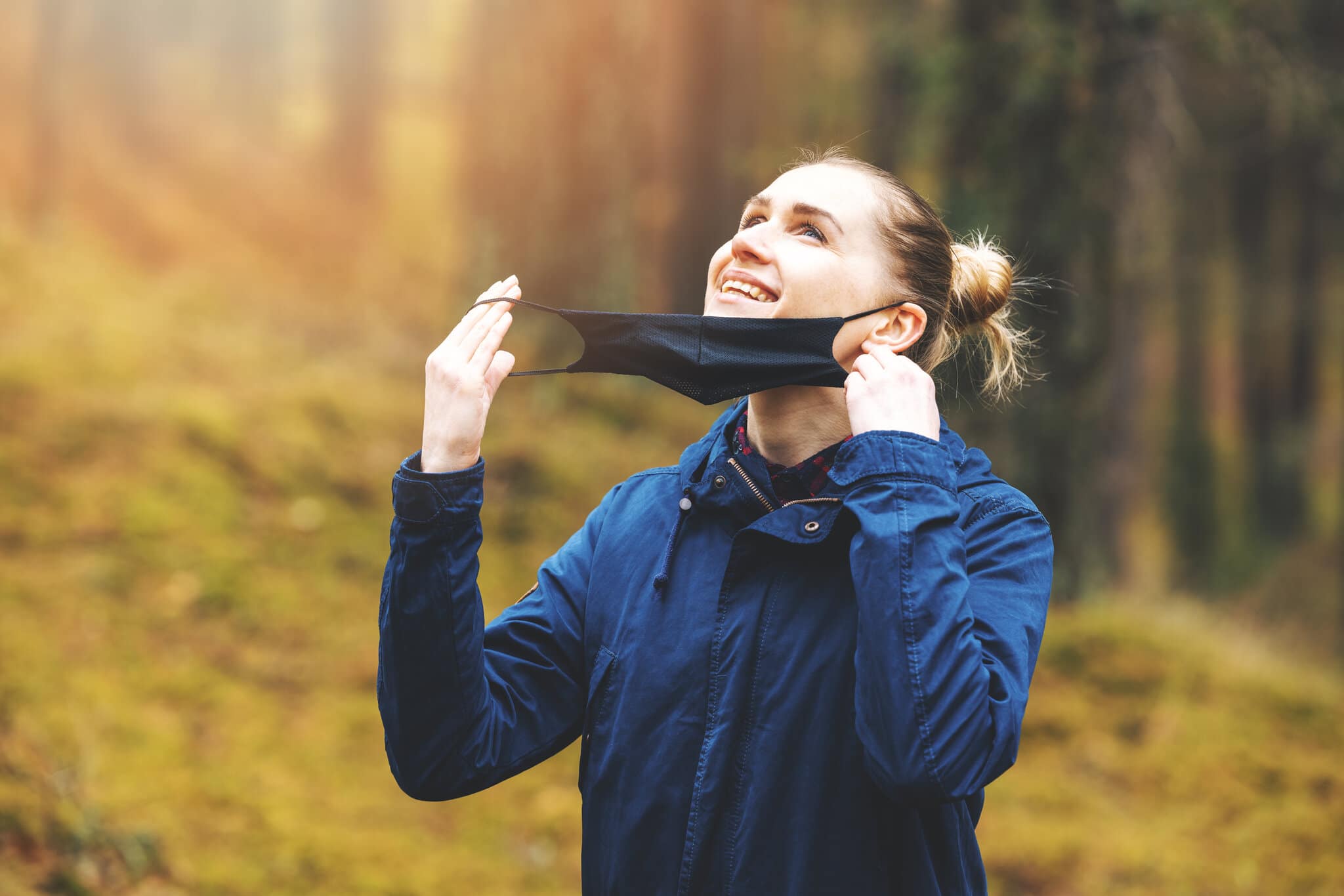 Young, smiling woman holds her face mask while enjoying a walk in nature